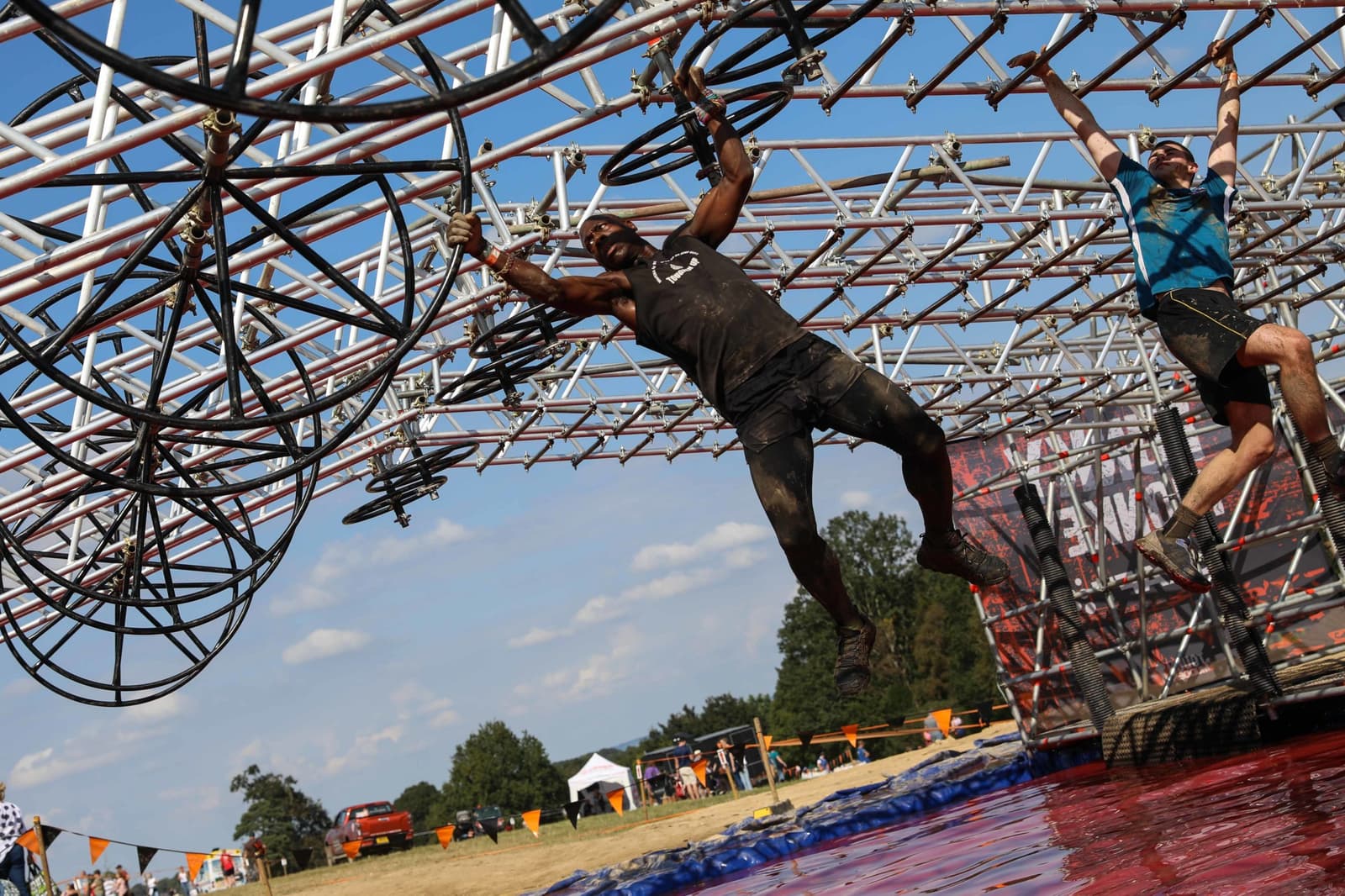 Two individuals participate in an obstacle course, hanging and traversing using metal rings. The man in the foreground is reaching forward while the other participant swings behind him. The ground below is red and muddy. Trees and a clear sky are visible in the background.