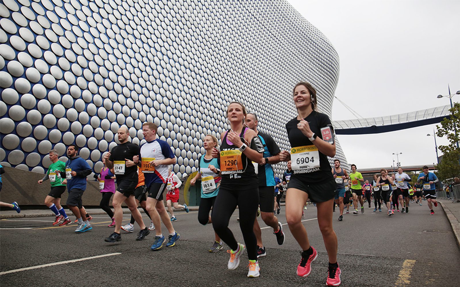 A diverse group of runners participating in a race, running on a city street in front of a modern building with a circular, textured facade. Some runners wear race bibs and colorful athletic gear, and the atmosphere appears lively and energetic.