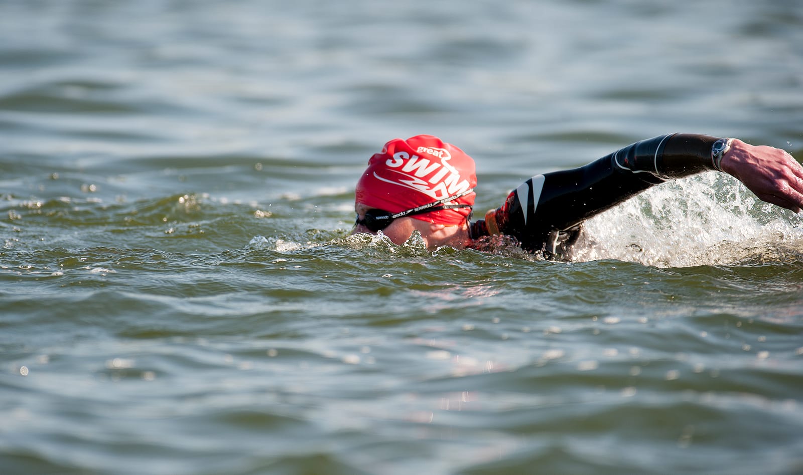 A swimmer wearing a red swim cap and black wetsuit is performing a freestyle stroke in open water. The cap has the word "SWIM" printed on it. The swimmer is partially submerged, with their face mostly in the water and their arm extended forward.
