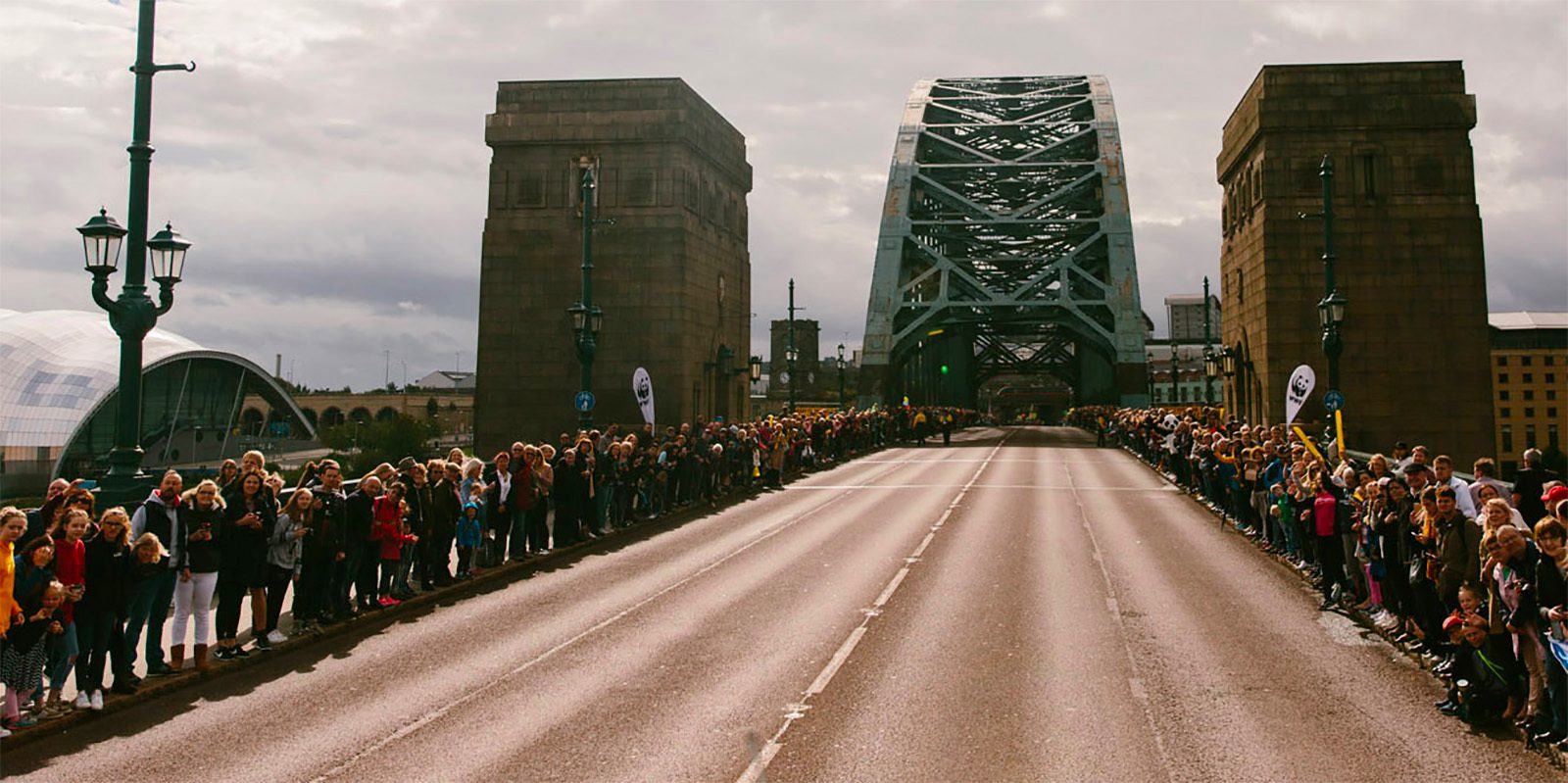 A large crowd of people is gathered on both sides of a road that leads to a towering bridge with steel arches and stone pillars. The sky is overcast, and the atmosphere suggests an awaiting event or parade.