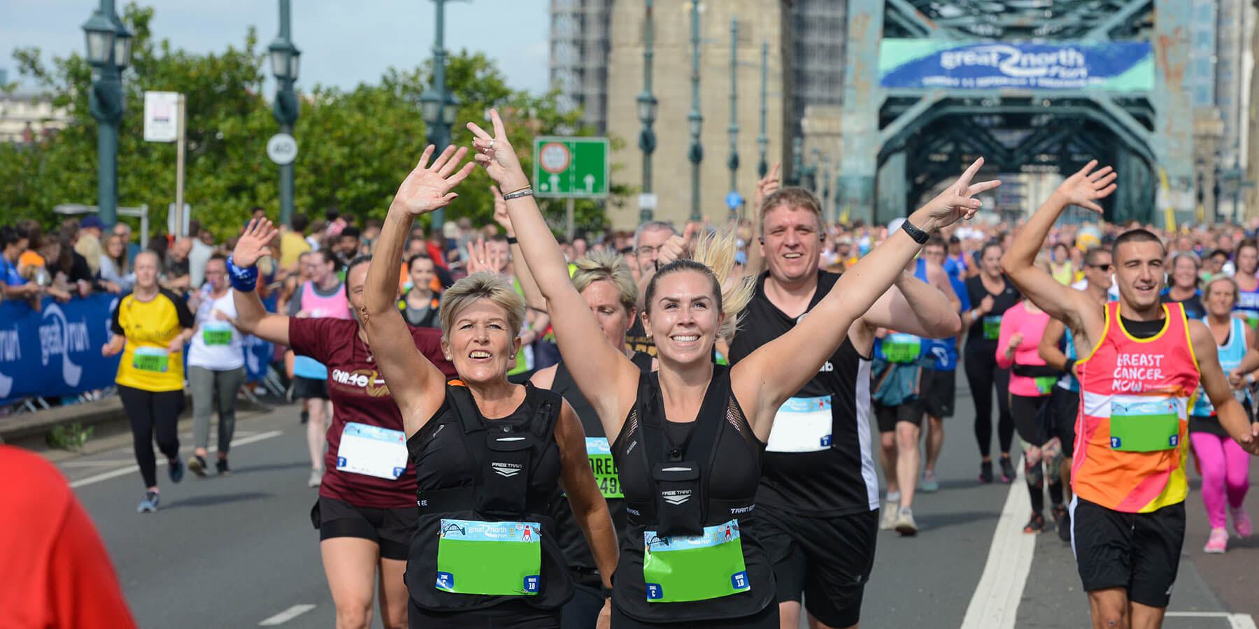 A large group of runners are participating in a marathon, smiling and waving as they run over a bridge. Many are wearing numbered bibs and colorful athletic clothing. Spectators line the route, and a large banner is visible in the background.