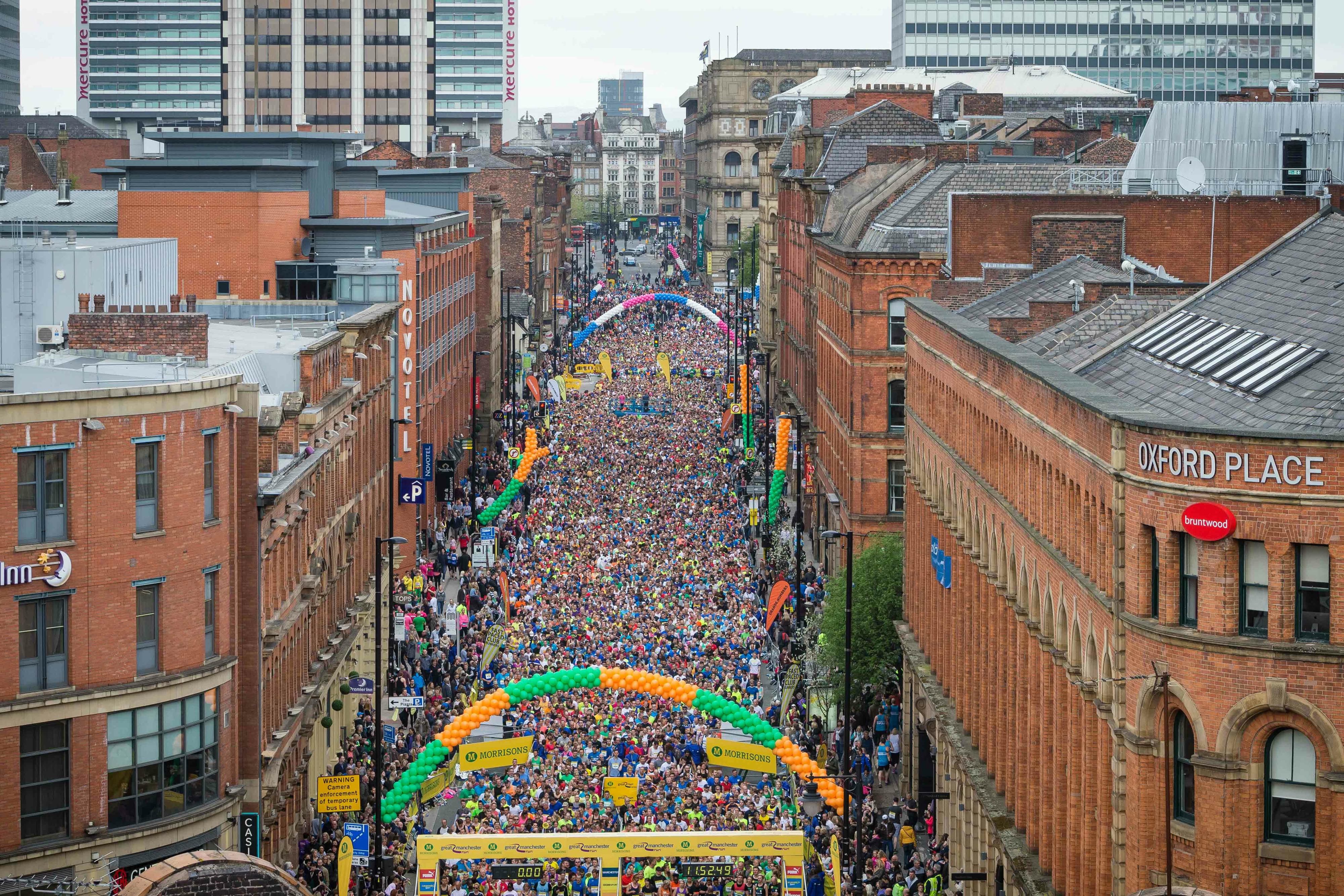 Aerial view of a crowded city street filled with participants in a running event. Brightly colored banners and balloons mark the start and finish lines. Tall buildings flanking the street, with a mix of older brick structures and modern high-rises in the background.