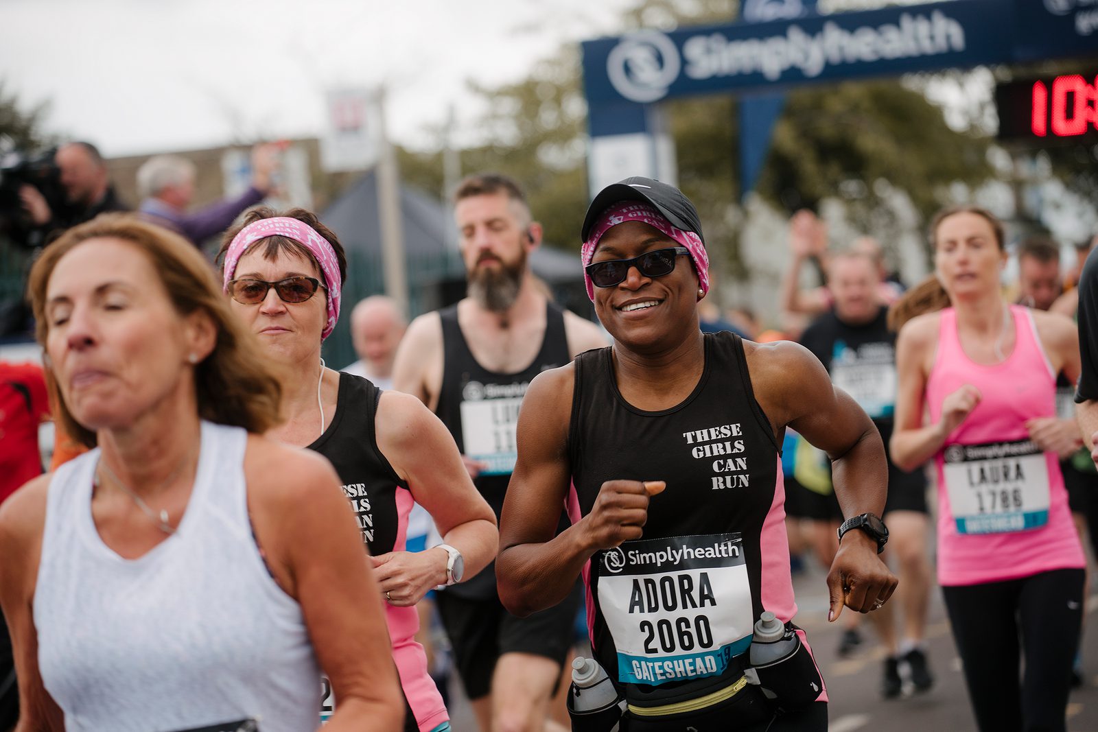 A group of enthusiastic runners participating in a race. The runners in the foreground wear black shirts with "THESE GIRLS CAN RUN" printed on them. One runner, sporting sunglasses and a wristwatch, smiles while running among the crowd. The race banner is visible in the background.