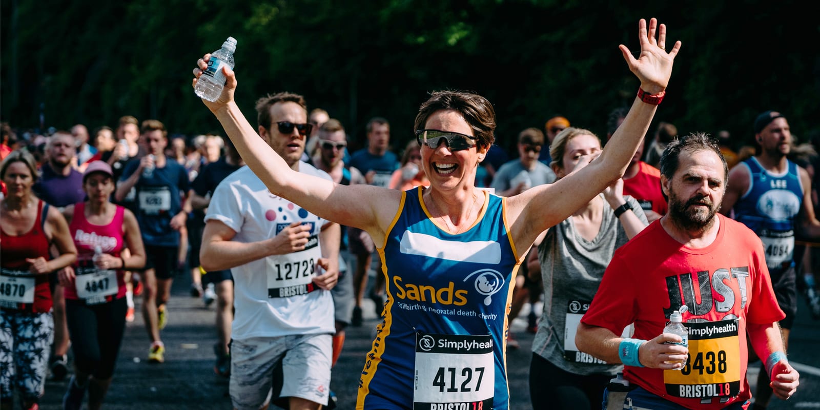A large group of marathon runners are on a road. A woman near the front, wearing sunglasses and a running vest labeled "Sands" and number 4127, raises her arms in victory while holding a water bottle. Other runners are around her, with some wearing numbered bibs.