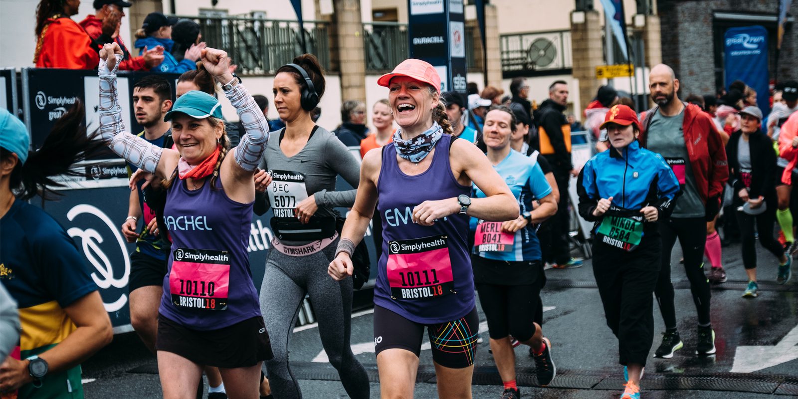 A group of runners participating in a race pass along a city street. Two women in the foreground, wearing numbered bibs and athletic gear, smile and raise their arms in celebration. Other runners, some wearing headphones and colorful outfits, follow closely behind.