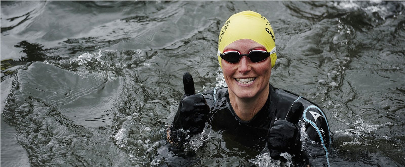 A person wearing a yellow swim cap, goggles, and a wetsuit is swimming in open water and giving two thumbs up while smiling. The water appears dark and rippling around them.