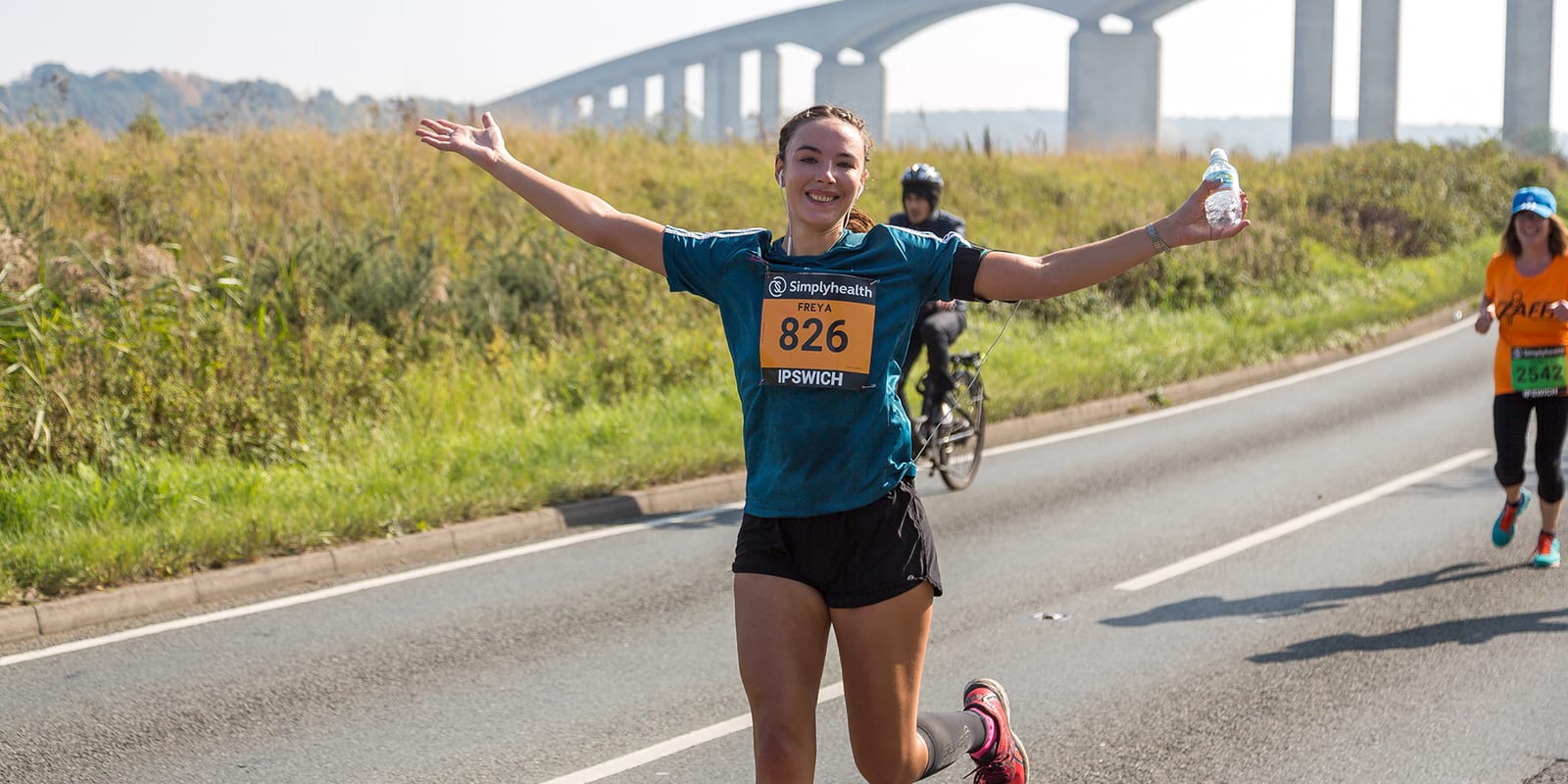 A runner wearing a blue T-shirt, black shorts, and race bib number 826, smiles and raises her arms while holding a water bottle. She runs on a paved road with a cyclist and another runner in the background. A bridge is visible behind them, and the scene is under clear skies.