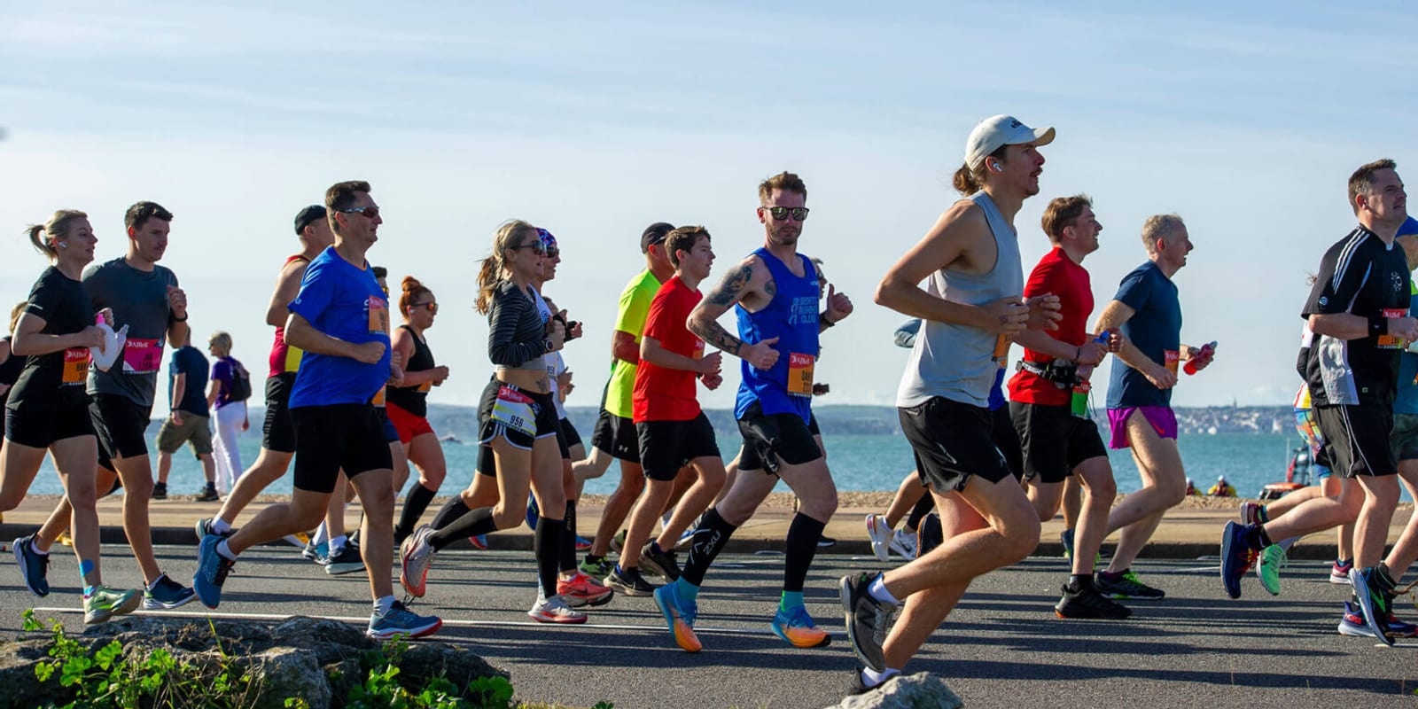A group of runners wearing athletic gear participate in a road race along a seaside route on a sunny day, with the ocean and blue sky visible in the background.