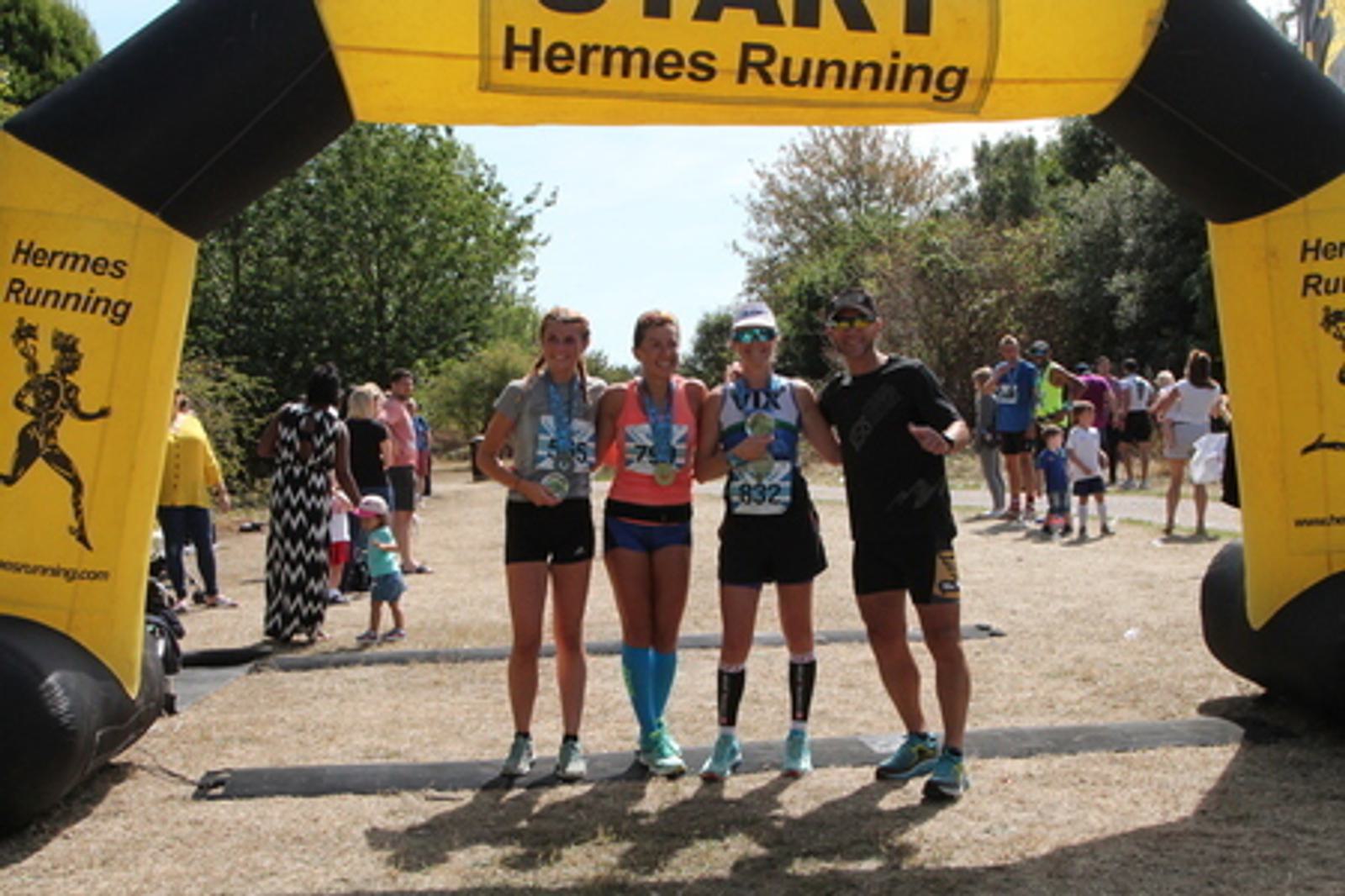 Four runners stand together under a yellow inflatable finish line arch labeled "Hermes Running." They are smiling and posing for a photo, with three holding trophies. Other participants and spectators can be seen in the background in a park setting.