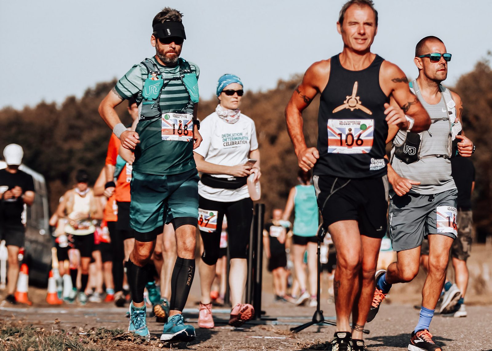 A group of runners participating in a race, wearing athletic gear and race bibs, moving along an outdoor course on a sunny day with trees in the background.