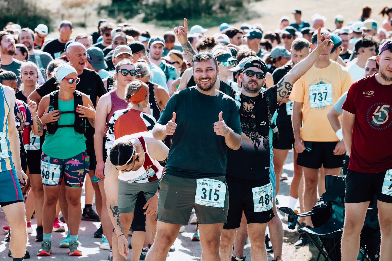 A group of runners gather at the start line of a race. One man in the center, wearing a numbered bib and shorts, smiles and gives two thumbs up. Other participants are chatting, stretching, or preparing to run.