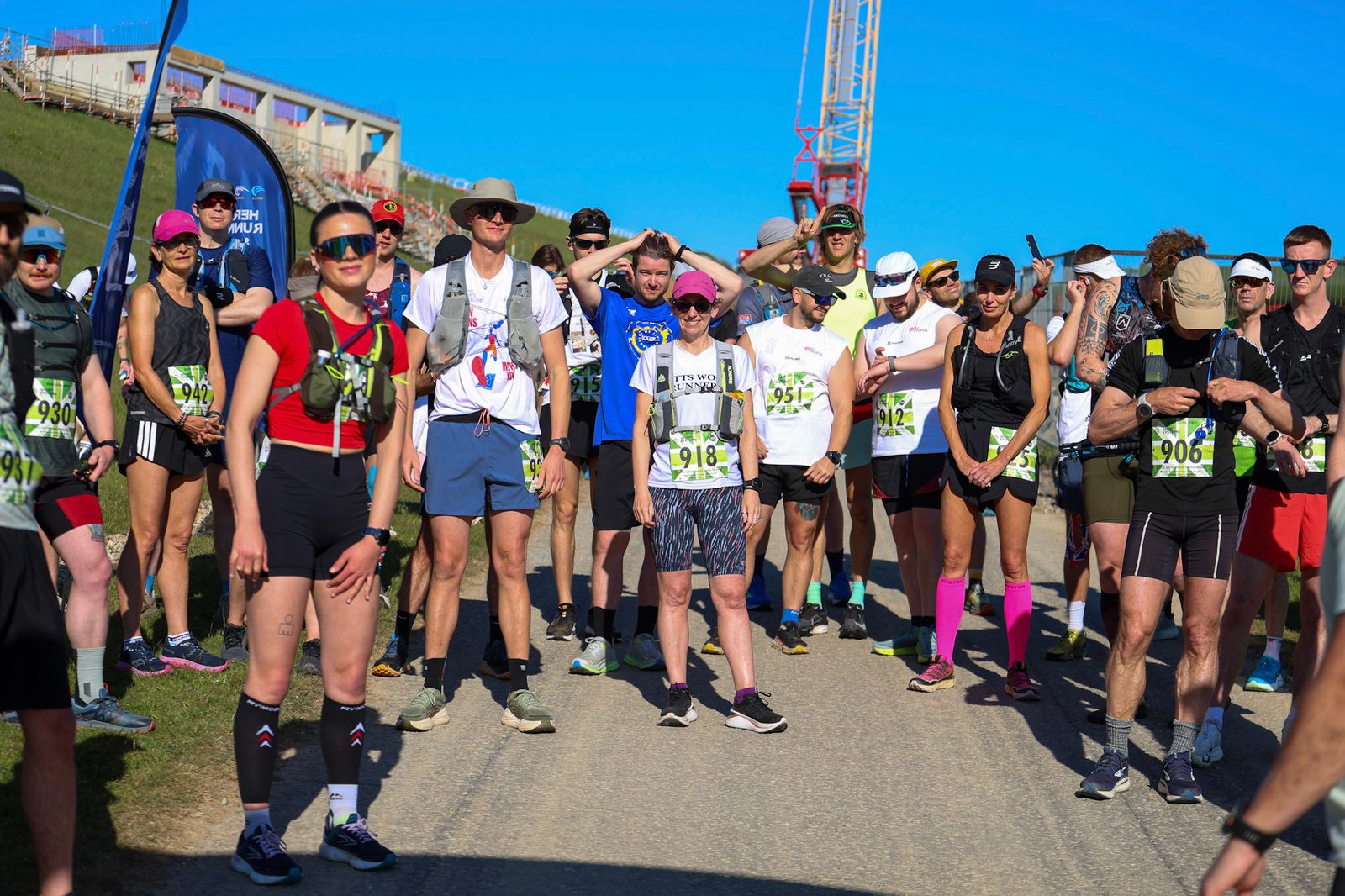 A group of runners wearing numbered bibs and athletic gear stand together on a sunny road, smiling and preparing for a race. Green hills and clear blue sky are in the background.