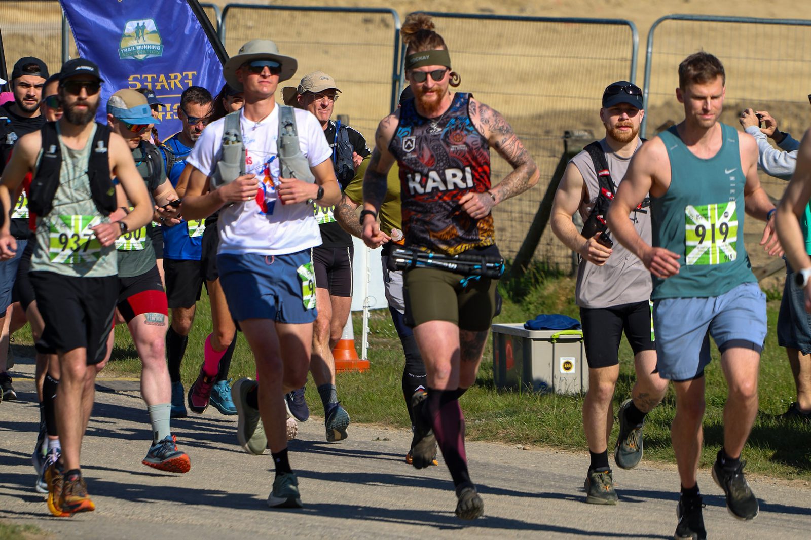 A group of men in athletic gear begin running a race outdoors on a sunny day, some wearing hats and sunglasses, with race numbers on their shirts and a "START" banner visible in the background.