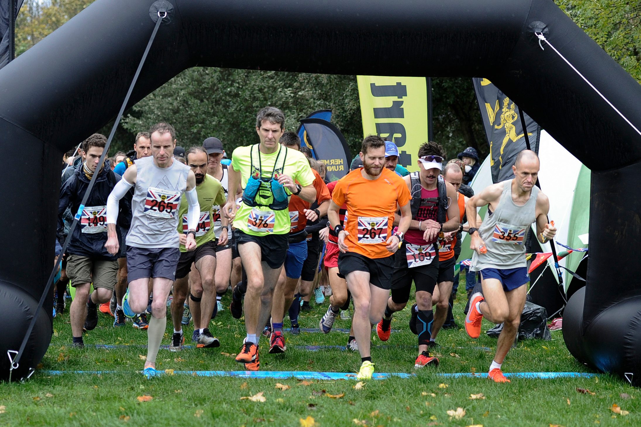A group of runners, wearing race bibs, start their race under an inflatable archway on a grassy field. Some participants are in colorful athletic gear. Spectators and volunteers can be seen in the background. The atmosphere is energetic and focused.