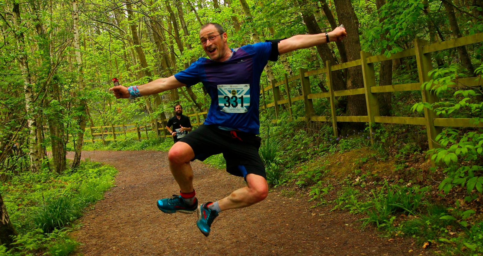 A man with a race bib numbered 331 joyfully jumps in mid-air on a forest trail. He is wearing a blue shirt, black shorts, and blue running shoes. The trail is surrounded by lush green foliage and a wooden fence is visible to the right. Another runner is in the background.