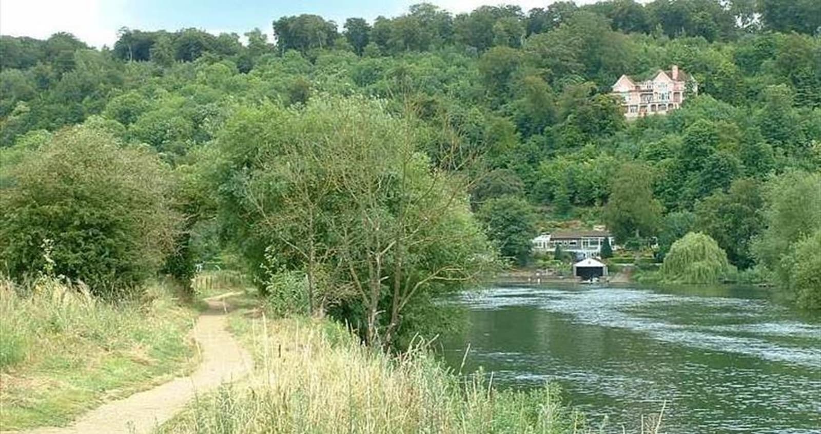 A scenic riverside path winds through lush greenery with a forested hill in the background. A large house sits atop the hill, overlooking the water. The calm river reflects the surrounding trees and vegetation, creating a peaceful natural setting.