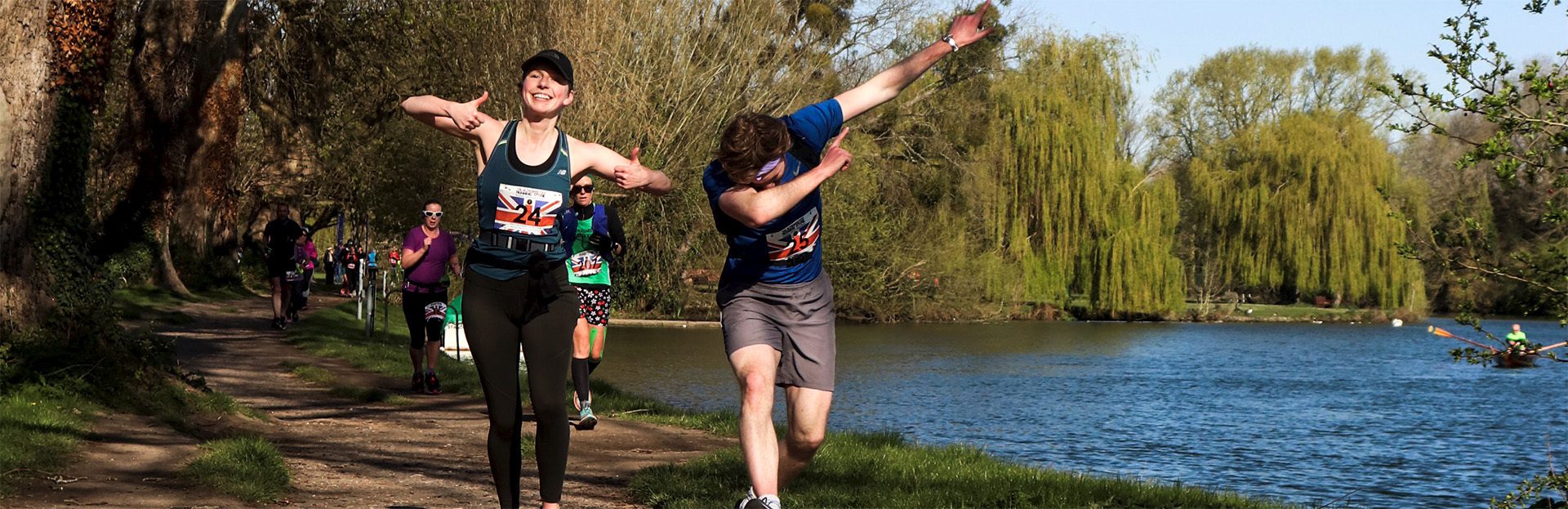 A man and a woman, both with race bibs, are running on a lakeside trail. The woman is smiling with her arms raised, while the man is performing a dab gesture. Other runners can be seen in the background along the path. The lake is surrounded by trees.