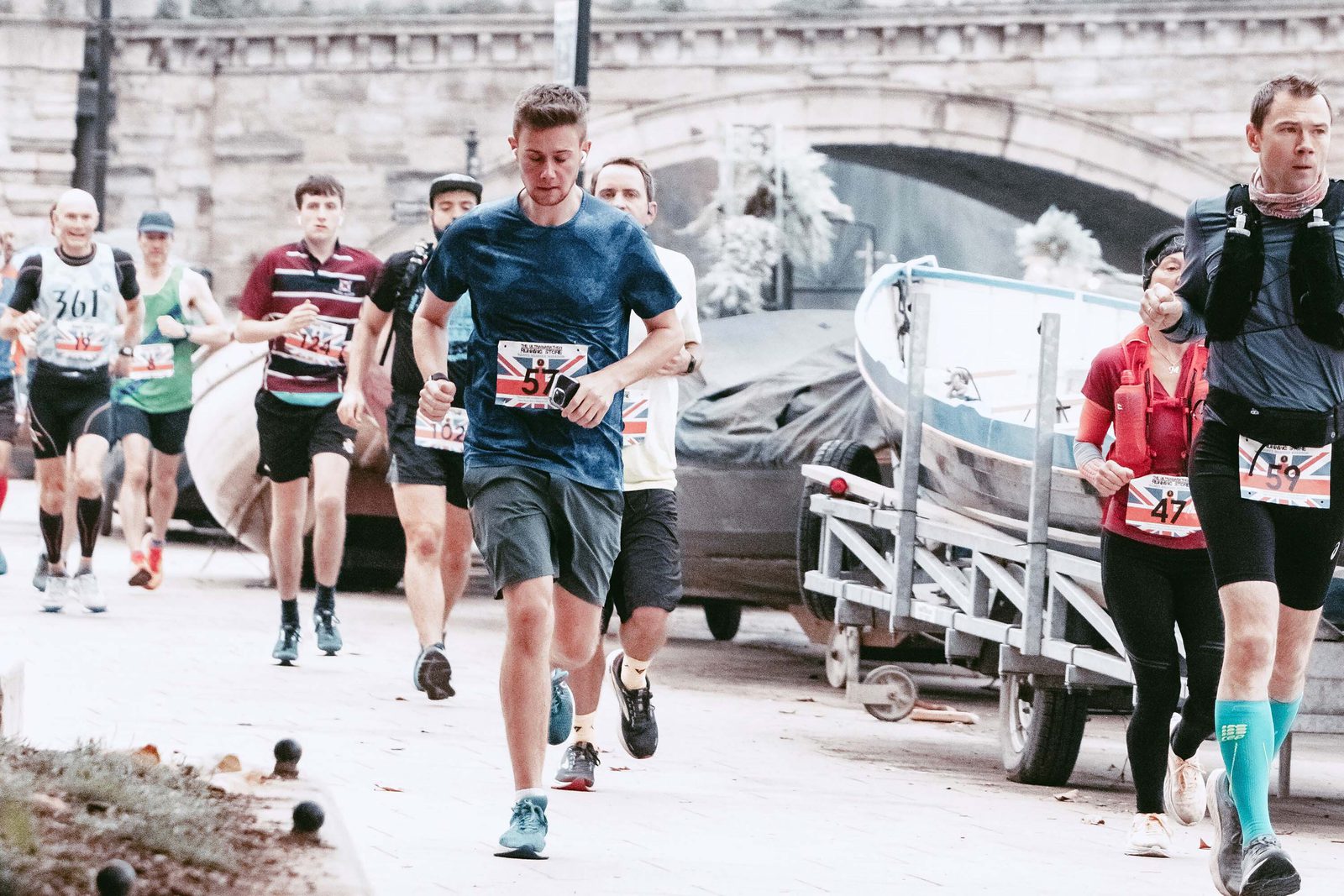 A group of runners participating in a race on a paved path beside a stone bridge. The leading runner, wearing a blue shirt and dark shorts, is closely followed by others in various athletic attire. Some spectators stand nearby. It's an overcast day.