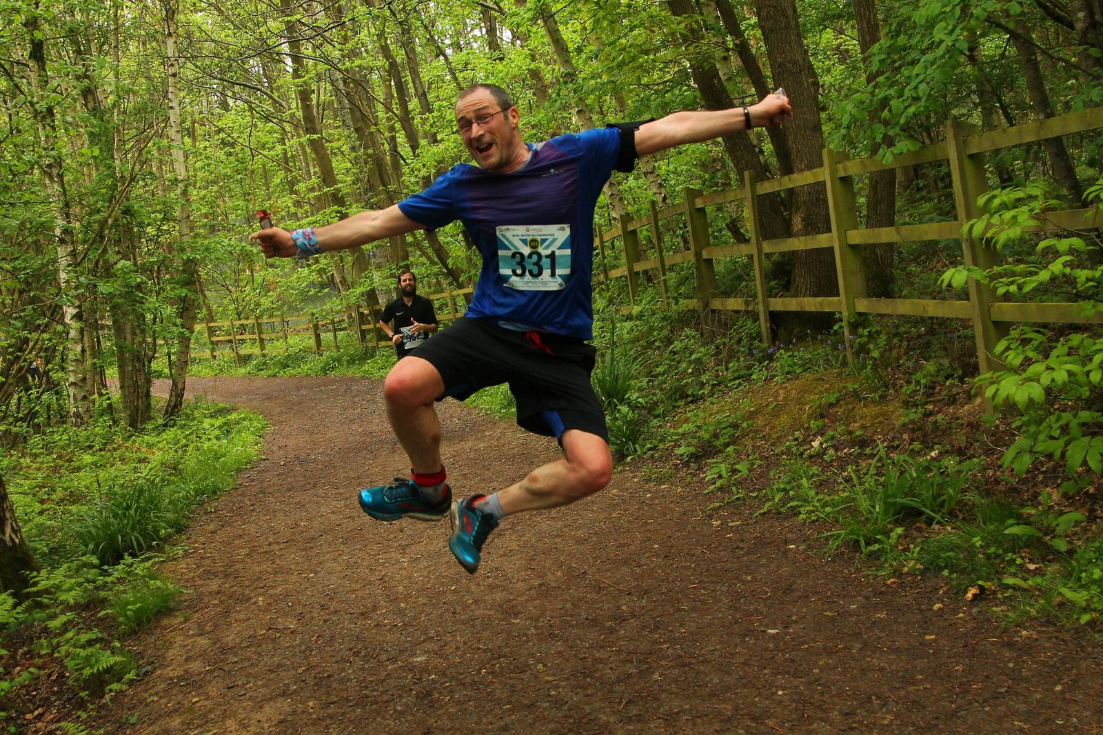 A runner with the number 331 on his chest is mid-air in a joyful leap on a forest path, lined with trees and a wooden fence. Another runner is seen in the background. The atmosphere is vibrant with lush green foliage.
