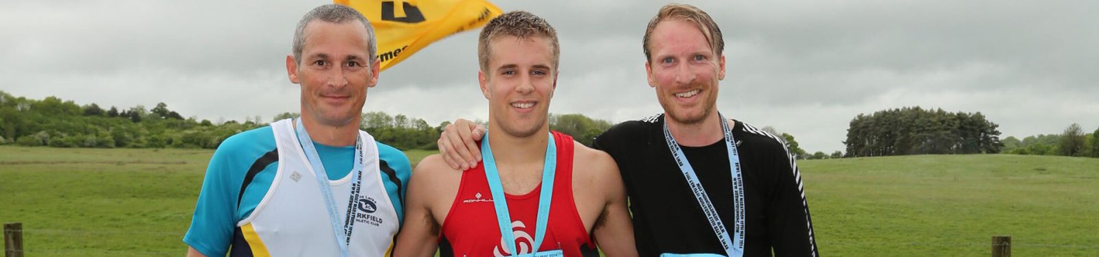 Three men stand side by side outdoors, each wearing a race bib and medals around their necks, smiling at the camera. The man on the left wears a white and blue jersey, the middle one a red jersey, and the one on the right a black jersey. A yellow flag flutters in the background.