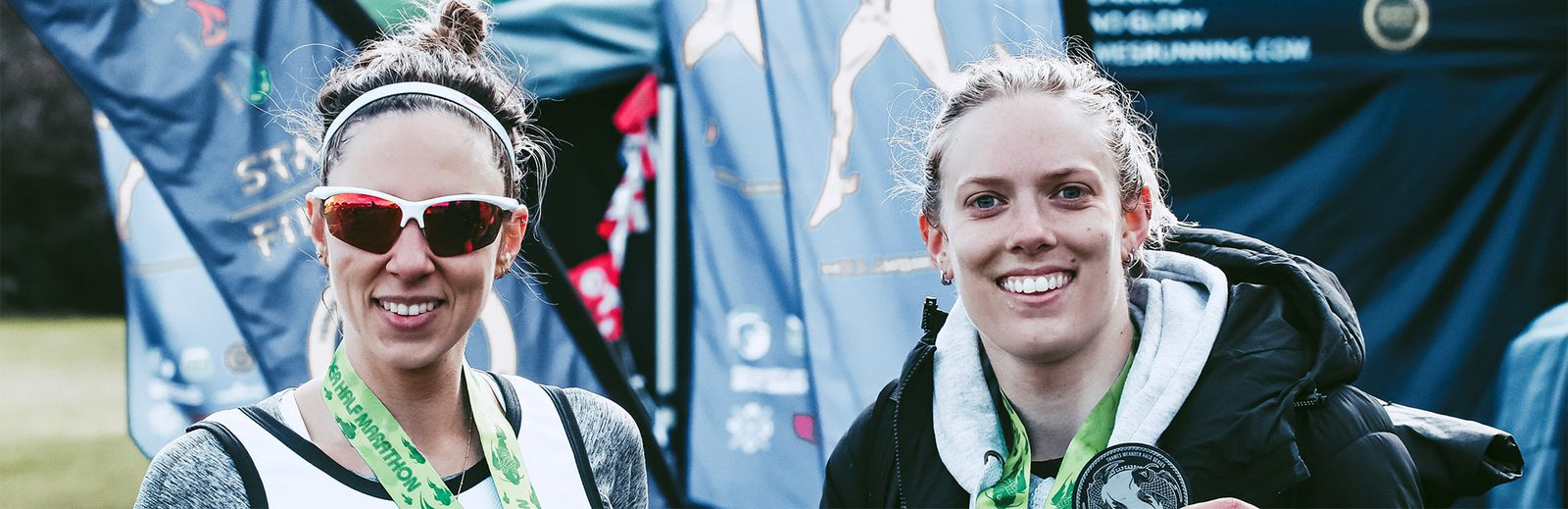 Two athletes pose for a photo after a race. They are smiling and wearing bright medals around their necks. The athlete on the left has sunglasses and a white headband, while the one on the right holds a grey medal and wears a black jacket. Various banners are visible in the background.