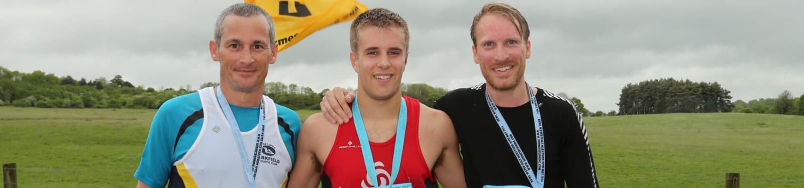 Three men stand side by side wearing medals, smiling at the camera. The man on the left wears a white and blue jersey, the man in the middle is in a red and blue tank top, and the man on the right is in a black shirt. They are outdoors with a green field in the background.