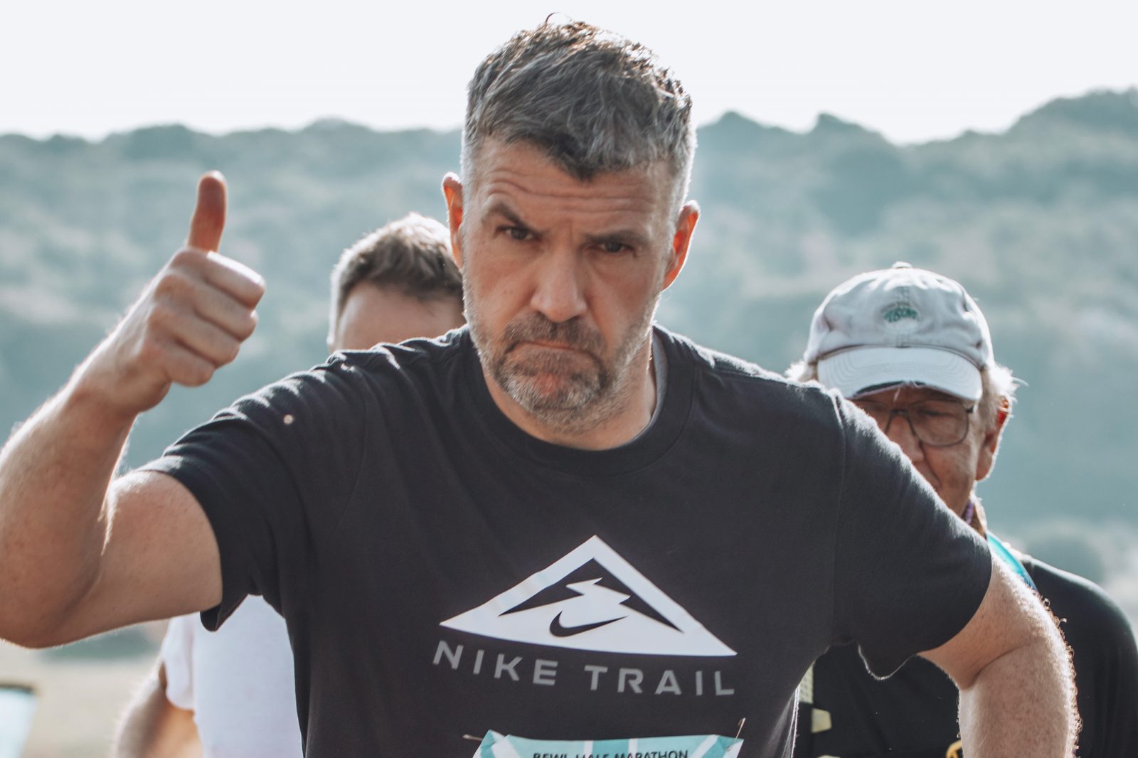 A man with gray hair and a beard gives a thumbs-up while participating in a marathon. He is wearing a black Nike Trail t-shirt. Other runners are visible in the background. The setting appears to be outdoors with a mountainous landscape.