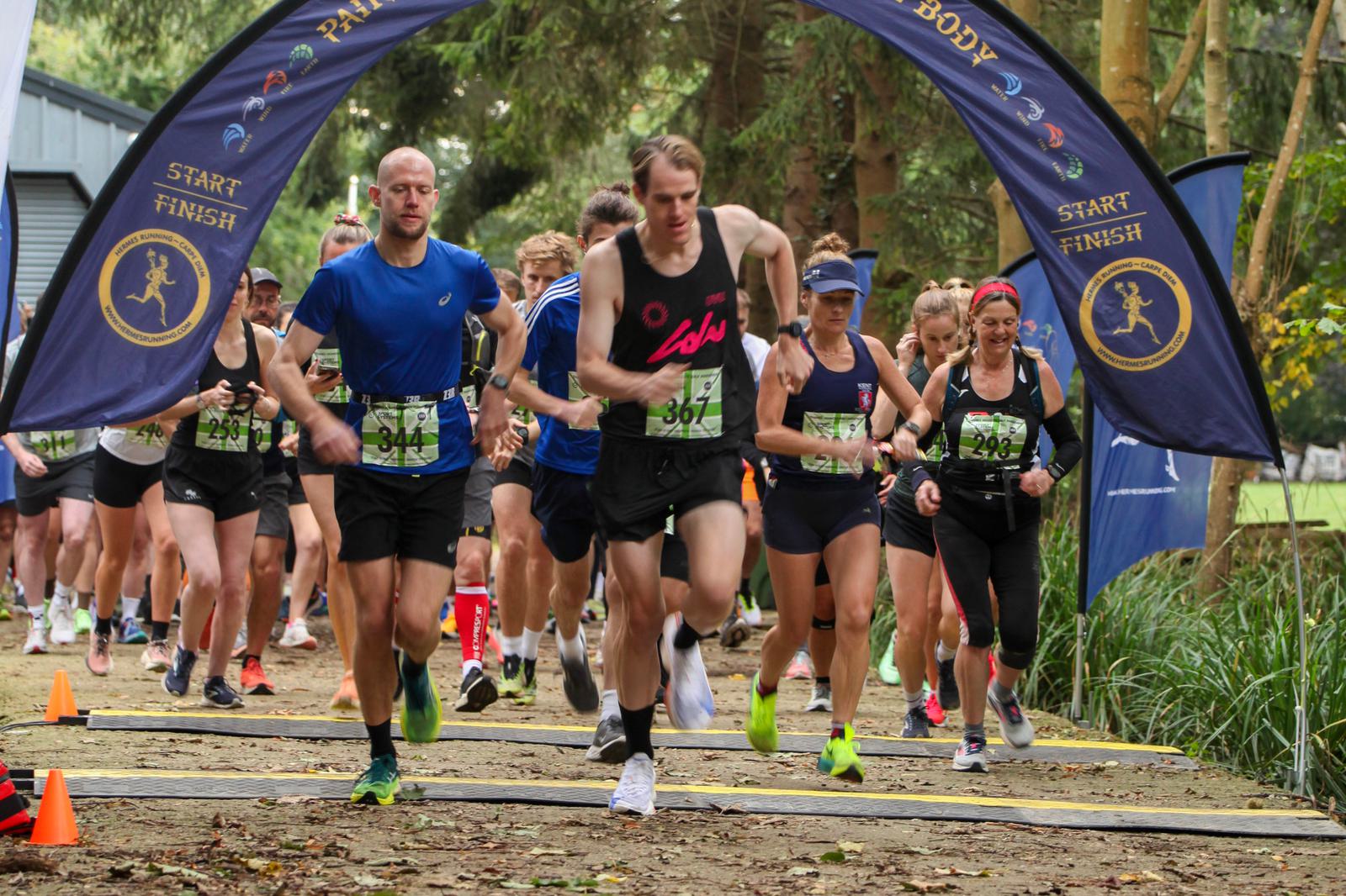 A group of runners, including men and women, is starting a race in a wooded area. They are passing under a blue arch with "Start" and "Finish" written on it. The scene is energetic, with participants wearing race bibs.