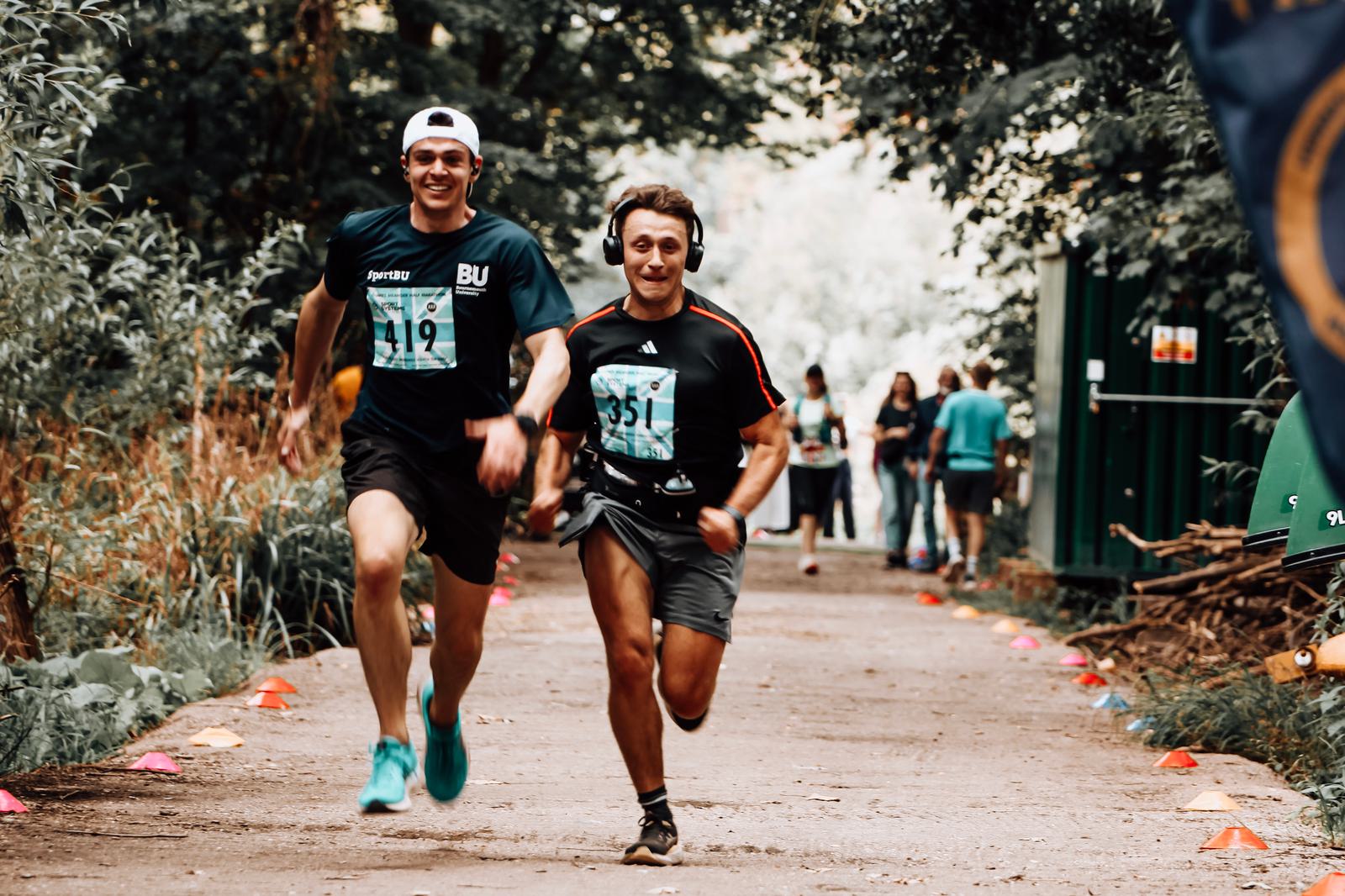 Two men running energetically in a race on a wooded path. Both are smiling, wearing sports attire with race numbers. The path is lined with small cones, and other participants can be seen in the background.
