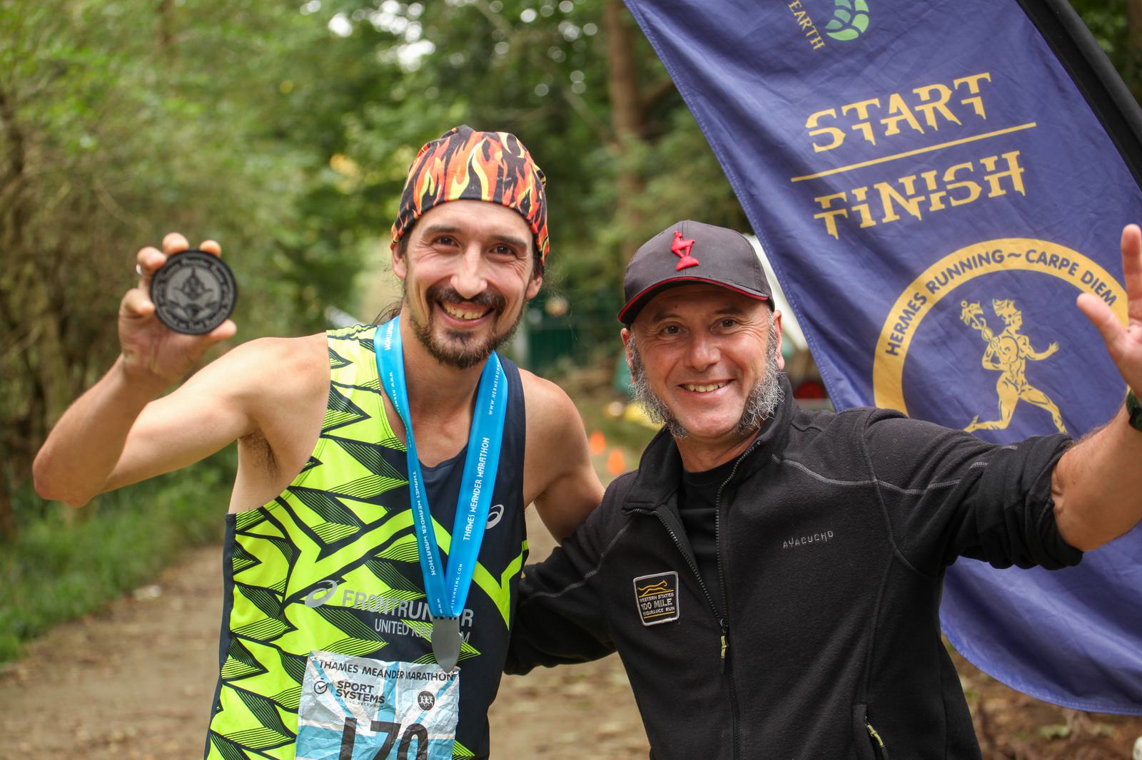 Two men are smiling and posing at a race finish. One holds a medal, wearing a bright running outfit and flame-patterned headscarf. The other man is in a dark jacket and cap. A "Start/Finish" banner is visible in the background.