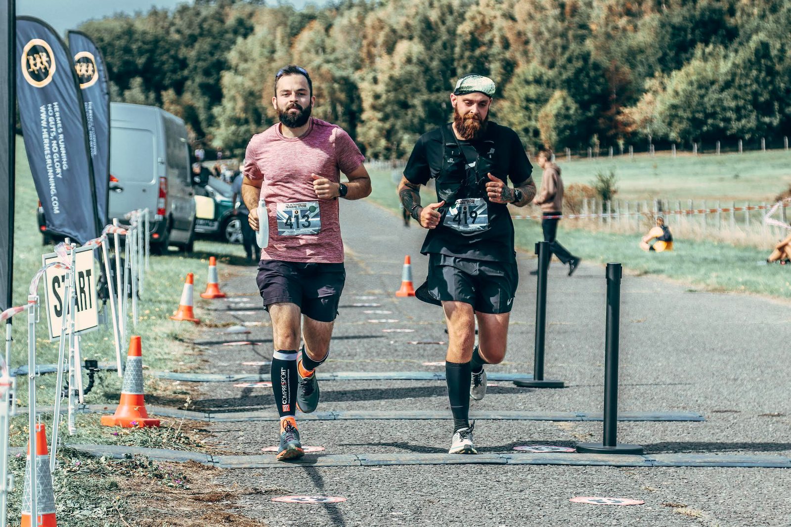 Two male runners with race bibs run side by side on a paved road during a race, passing the start line. There are safety cones, banners, and trees in the background.
