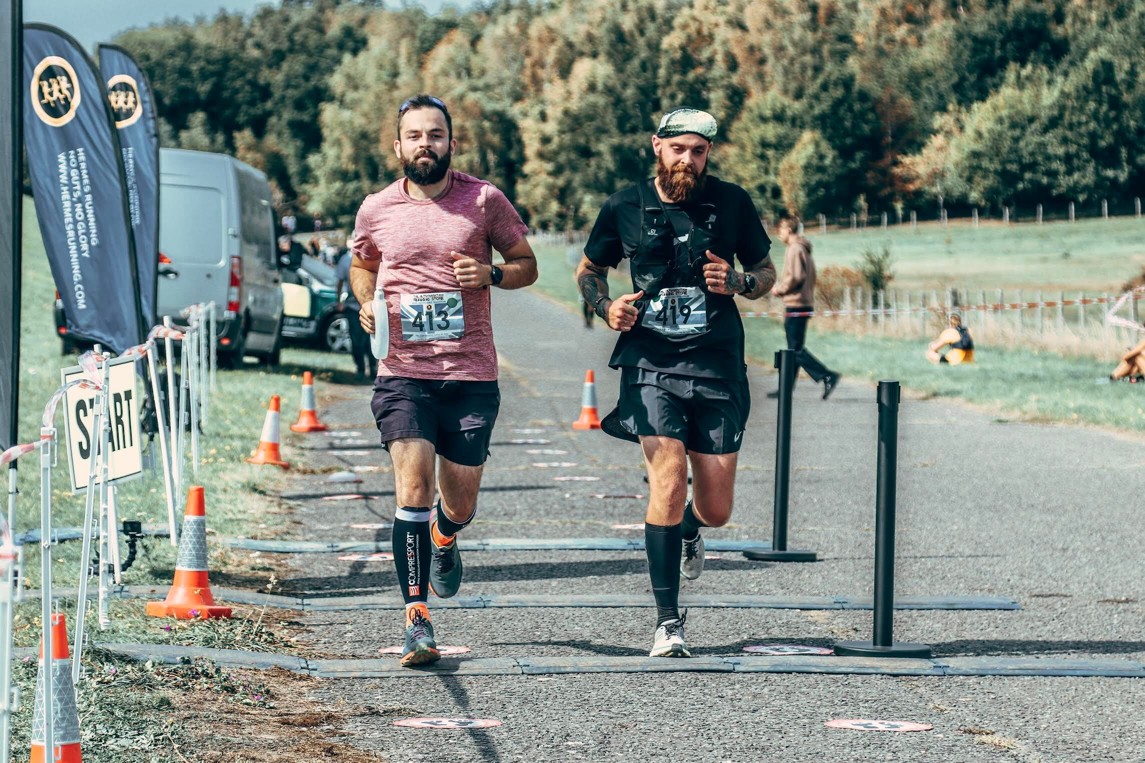 Two male runners with race bibs run side by side on a paved road during a race, passing the start line. There are safety cones, banners, and trees in the background.