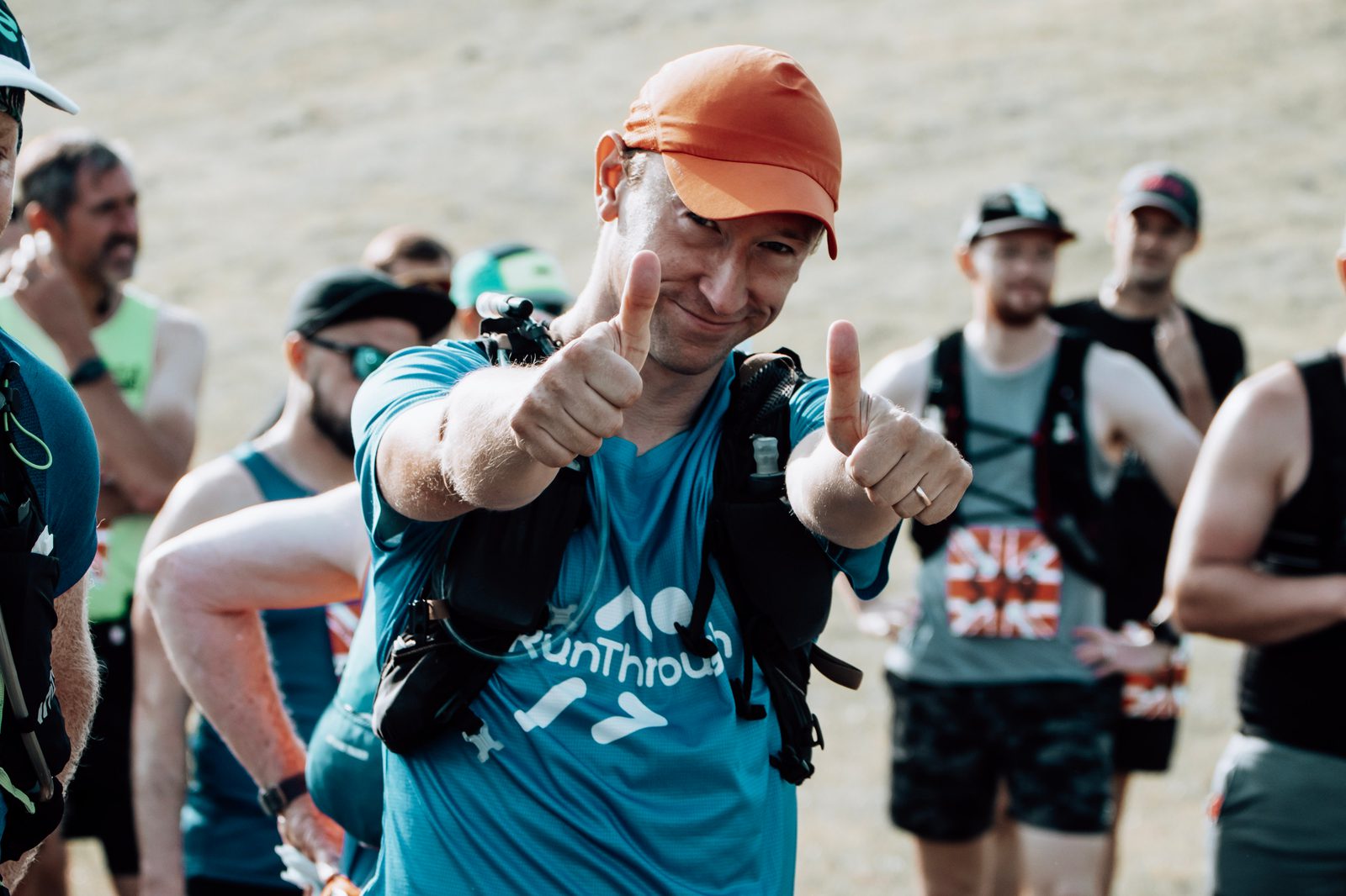 A smiling runner in a blue shirt and orange cap gives two thumbs up at an outdoor event, surrounded by other participants preparing to race.