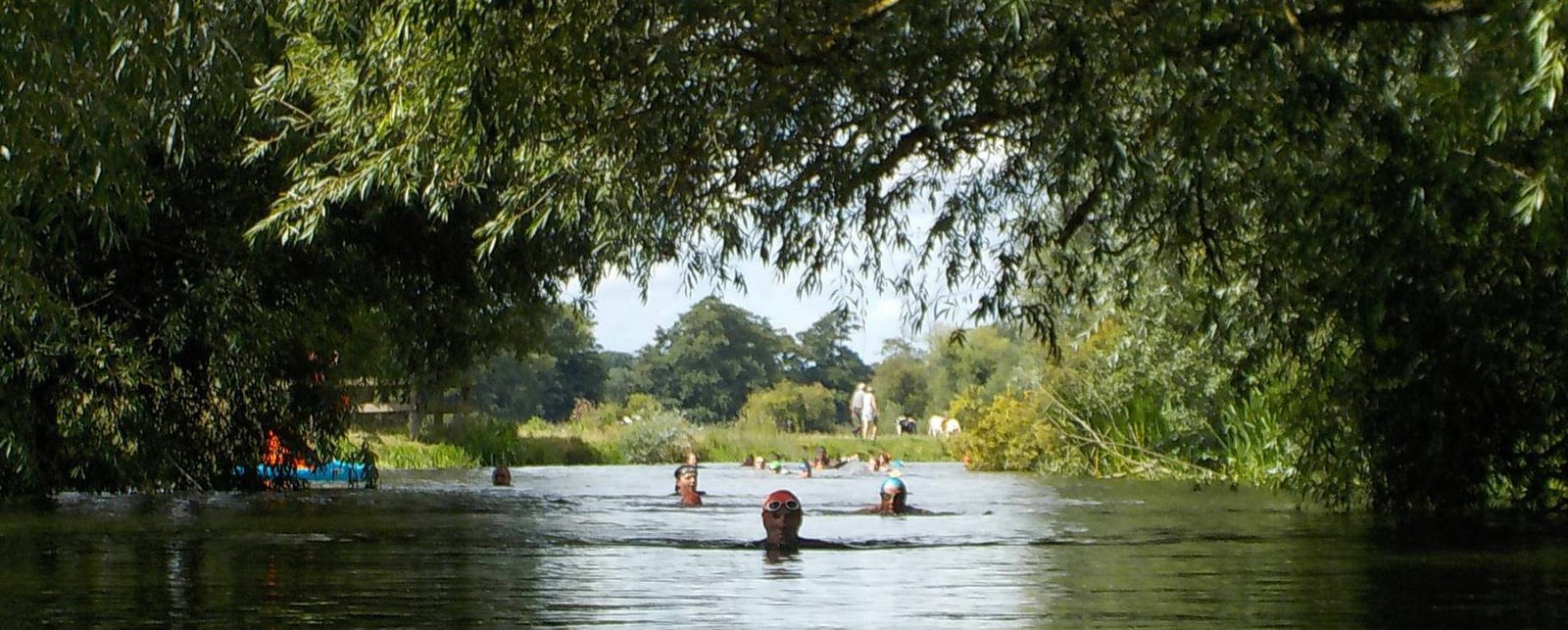 A group of people swim in a river surrounded by lush greenery on both sides. One person in the foreground is wearing goggles and a swimming cap. Trees arch over the water, creating a shaded area. A few people are visible in the background near the riverbank.