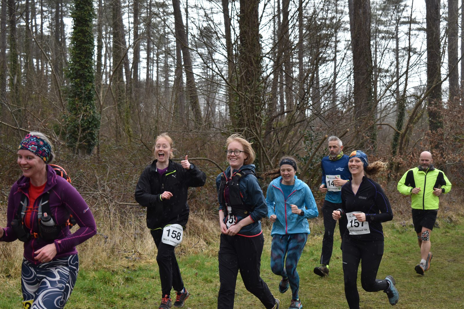 A group of runners, dressed in colorful athletic wear, jogging on a wooded trail. Some are smiling and giving thumbs up. They have race bibs pinned to their clothing with numbers such as 158, 151, and 184. The background shows tall trees and a forested area.