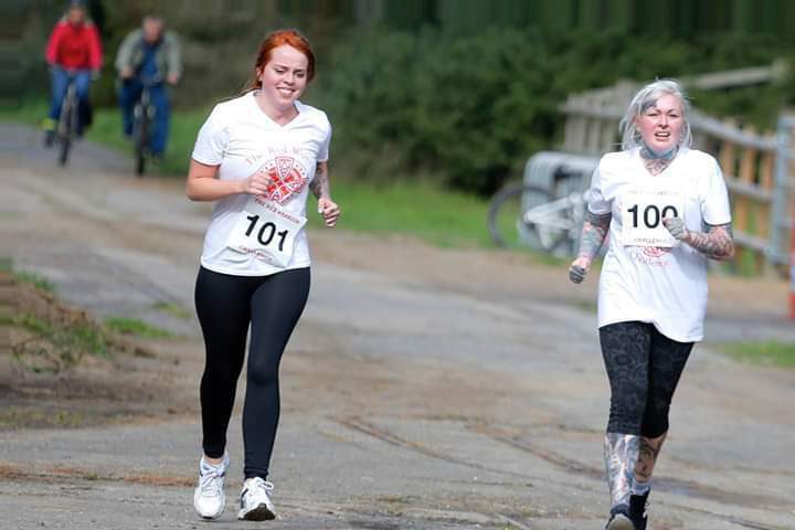 Two people are running on a paved path in a race. Both are wearing white T-shirts with numbers 101 and 100. The person on the left has red hair and is wearing black leggings. The person on the right has gray hair and tattoos, and is wearing patterned leggings. Two cyclists are in the background.
