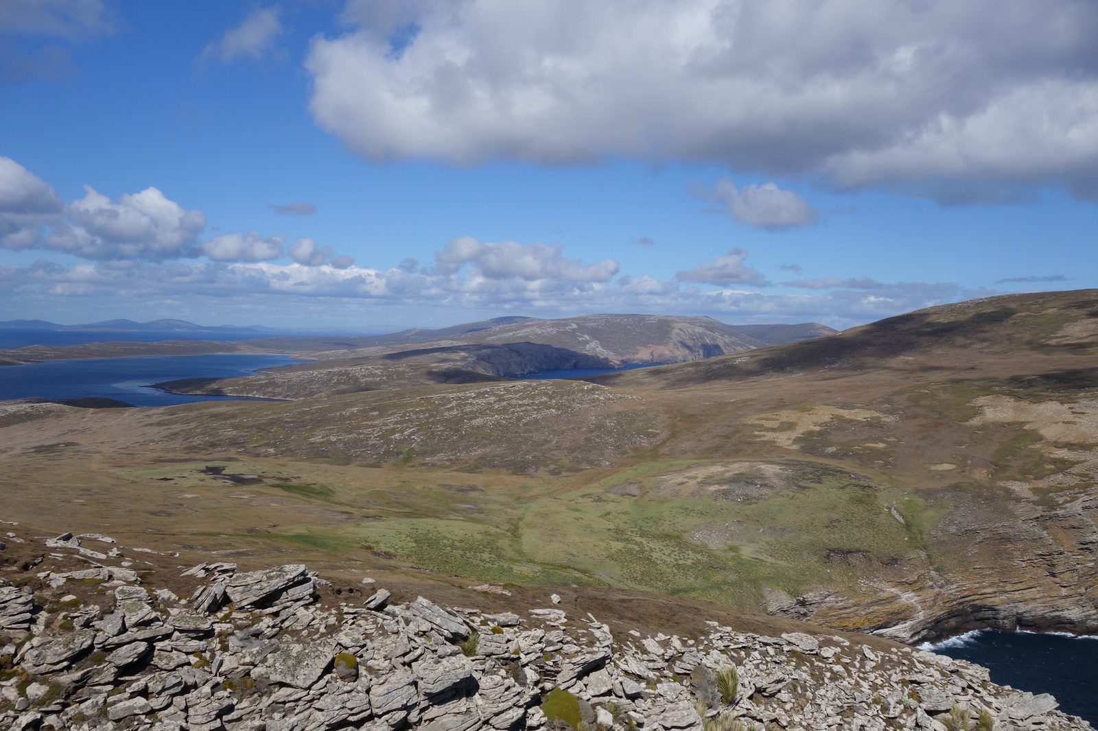 A wide-angle view of a rugged landscape with rocky terrain in the foreground. The middle ground shows a mixture of grassy and rocky areas, while rolling hills and distant mountains are beneath a blue sky with scattered clouds. A body of water is visible in the background.