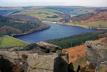 A scenic view of a large reservoir surrounded by green and brown rolling hills, with a bridge crossing over the water. Rocky outcrops are visible in the foreground, and the landscape extends into the distance under a partly cloudy sky.