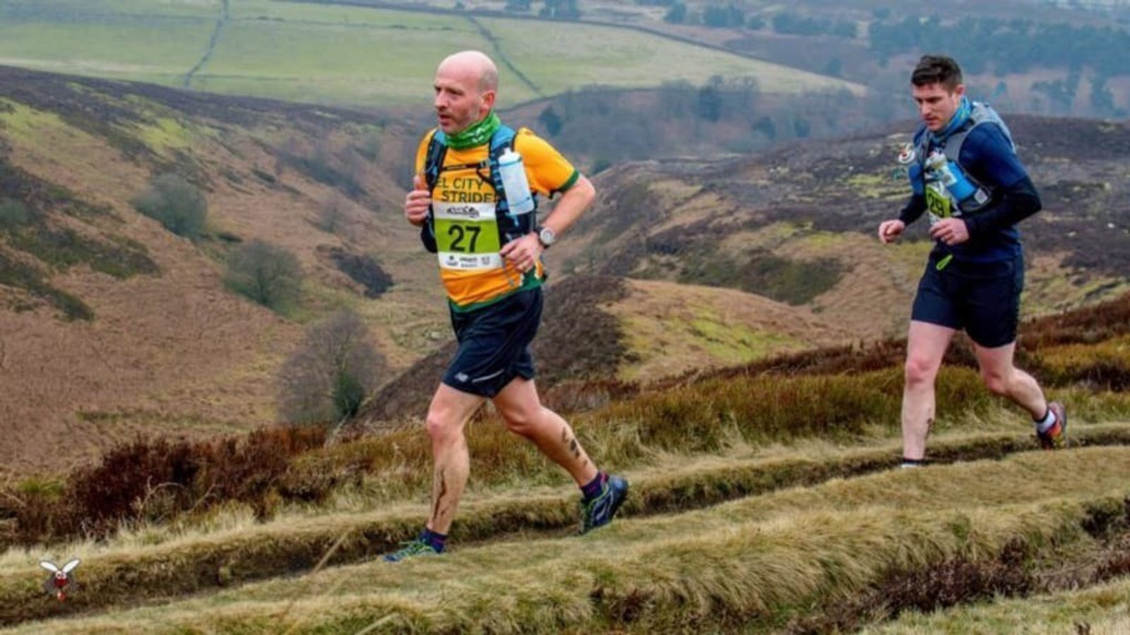 Two runners compete in an outdoor trail race on a hilly landscape. The leading runner wears a yellow and green jersey with the number 27, while the second runner wears a dark blue outfit with the number 115. The background features rolling hills and a cloudy sky.