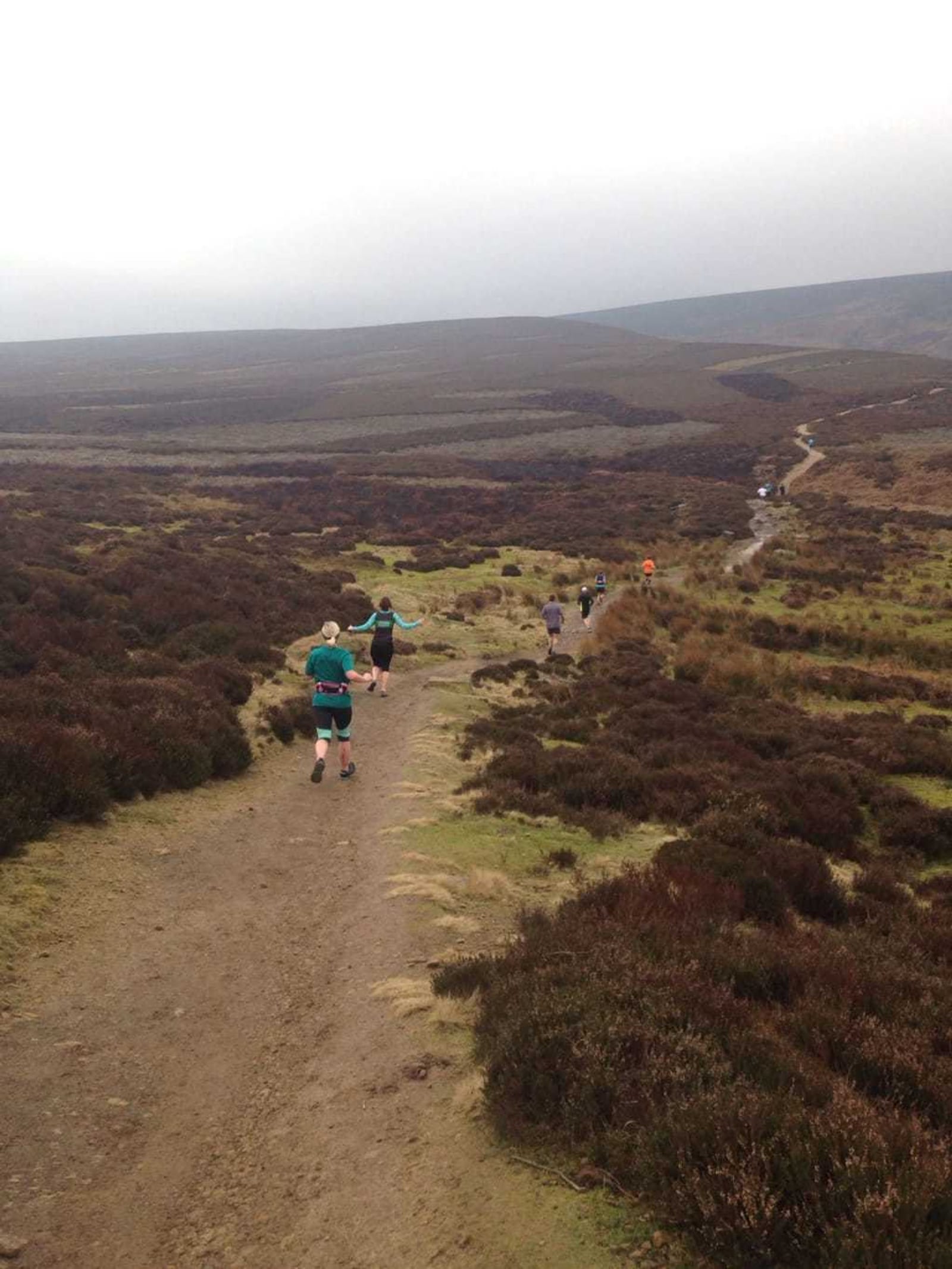 A group of people, mostly in athletic attire, are running on a trail through a vast, hilly moorland. The terrain is rugged with patches of grass and heath. The sky is overcast, and the landscape stretches far into the distance.