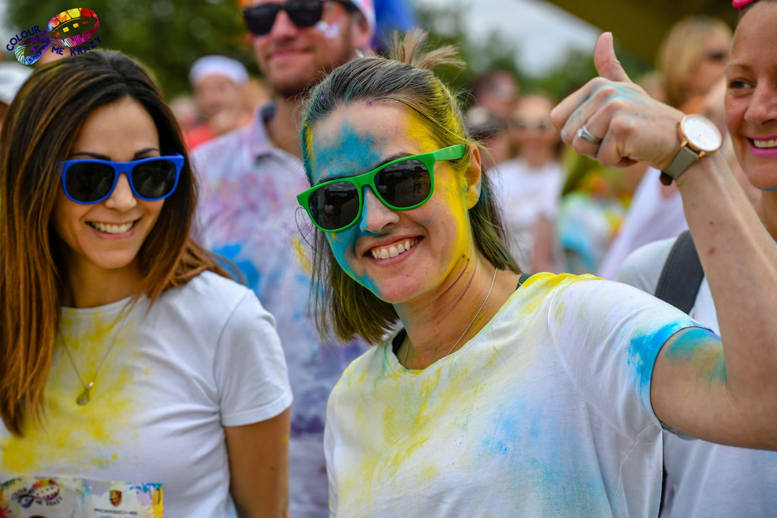 A woman wearing blue sunglasses and a white t-shirt is smiling and giving a thumbs-up, covered in colorful powder during a fun run event. She stands among other participants, some also wearing sunglasses and white t-shirts, with blurred outdoor scenery in the background.