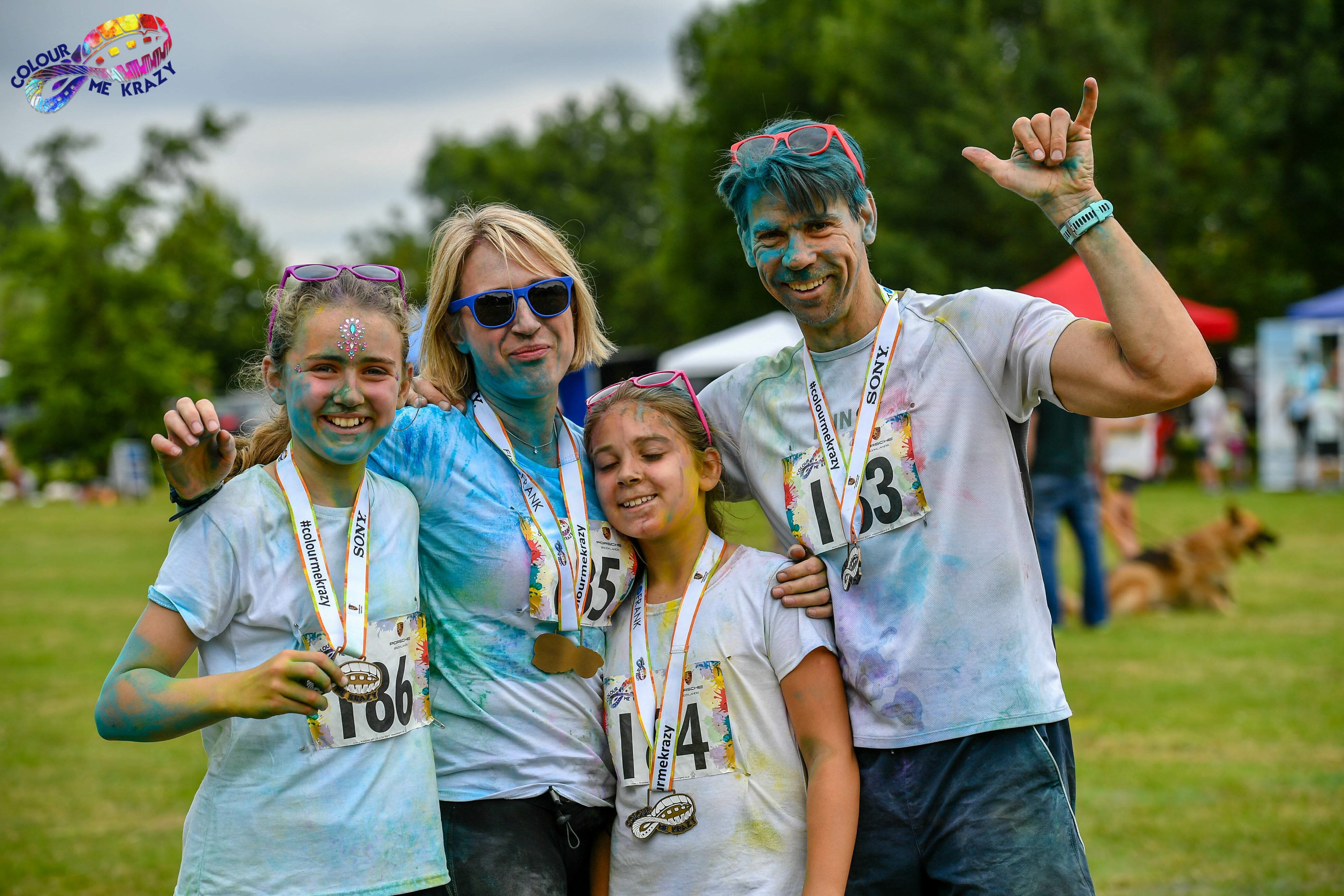 A family of four, wearing white shirts and medals, stands in a park covered in colorful powder. They are smiling, with the adults holding up their hands in celebratory poses. The father has blue hair, and the background features trees and tents.