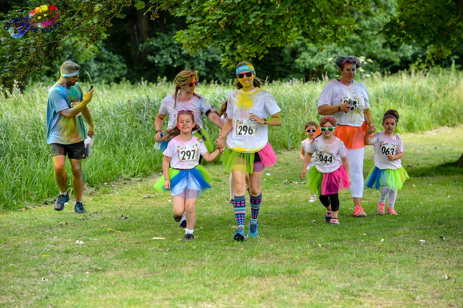 A group of people, including adults and children, participate in a colorful fun run or race. They are dressed in white shirts, vibrant tutus, and colorful socks, with numbers pinned to their shirts. Some have paint on their clothes and are smiling under leafy trees in a grassy area.