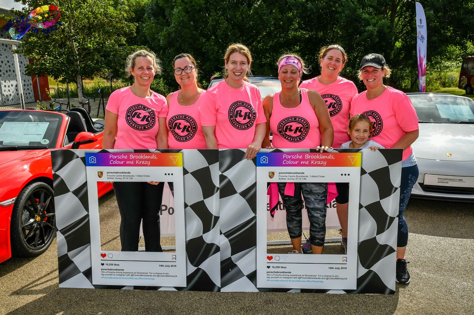 A group of seven smiling people, mostly women, wearing matching pink shirts stands in front of sports cars. They are holding two large Instagram photo frame props. Trees and a colorful logo are visible in the background.
