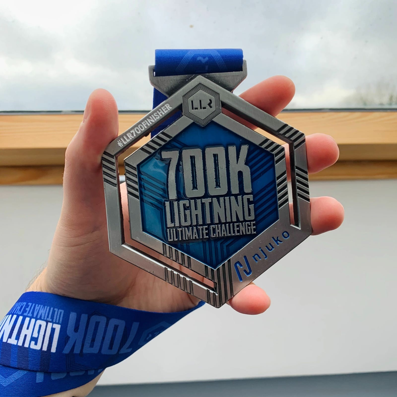 A person holds up a hexagonal medal with blue and metallic elements and the text "700k Lightning Ultimate Challenge" and "#LR700kfinisher". The medal is attached to a blue ribbon with "700k" and "Lightning Ultimate Challenge" printed on it.