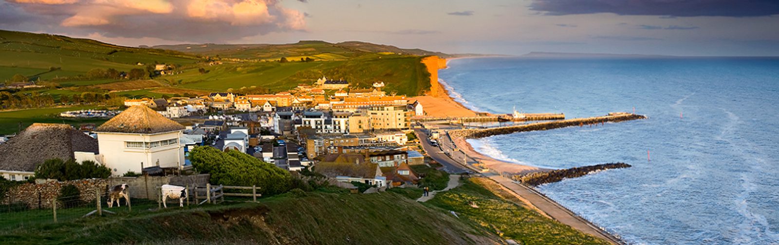 Panoramic view of a coastal village at sunset, featuring houses and buildings near the shoreline. The ocean waves meet a pebbled beach bordered by green hills. Wooden fences and livestock can be seen in the foreground, while the sky displays hues of orange and pink.