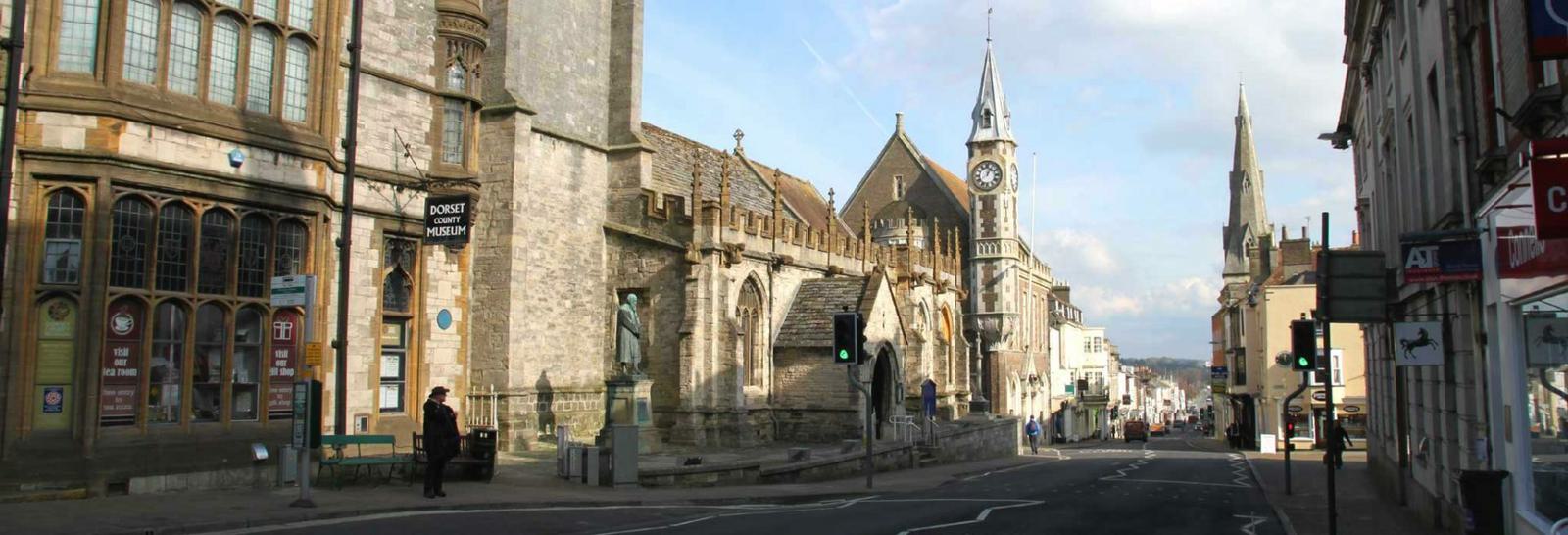 A street scene in a historic town featuring old stone buildings, a prominent clock tower, and a church. The road is lined with various structures, including shops and houses, under a partially cloudy sky. Few pedestrians are visible on the sidewalk.