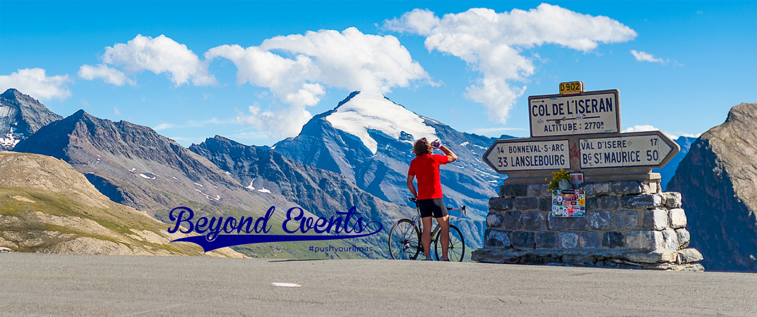 A person in a red jacket and black shorts stands beside a bicycle, taking a photo at the Col de l'Iseran mountain pass. Road signs indicate directions and distances to nearby locations. Snow-capped peaks and a clear blue sky form the scenic backdrop. Text reads "Beyond Events.