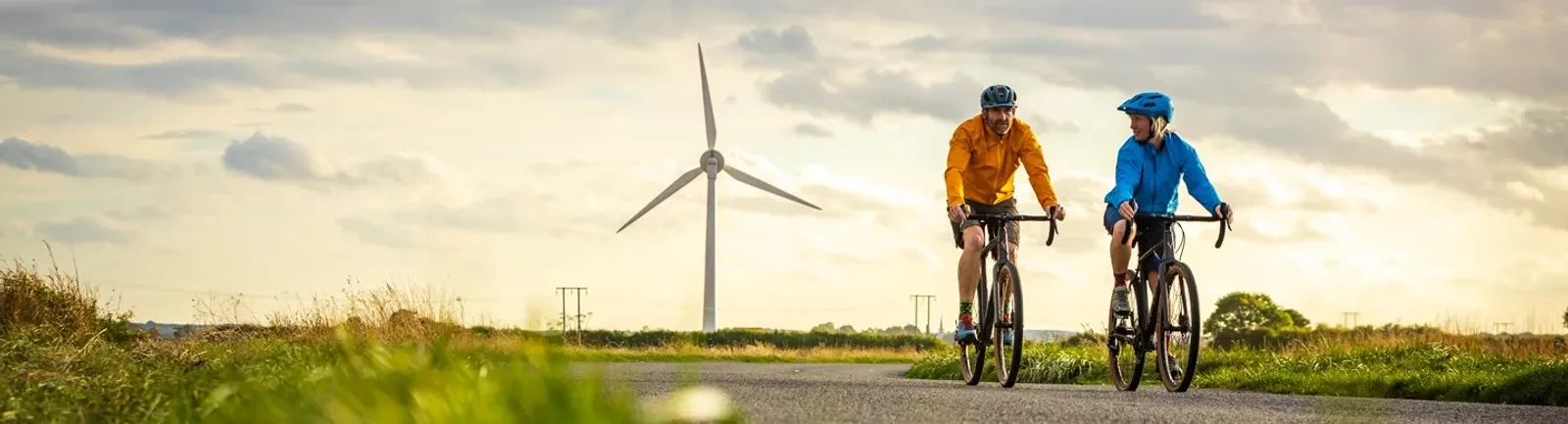 Two cyclists wearing helmets and jackets are riding on a rural path with a wind turbine visible in the background. The sky is partly cloudy, and the surrounding landscape is lush and green, creating a scenic and serene setting.