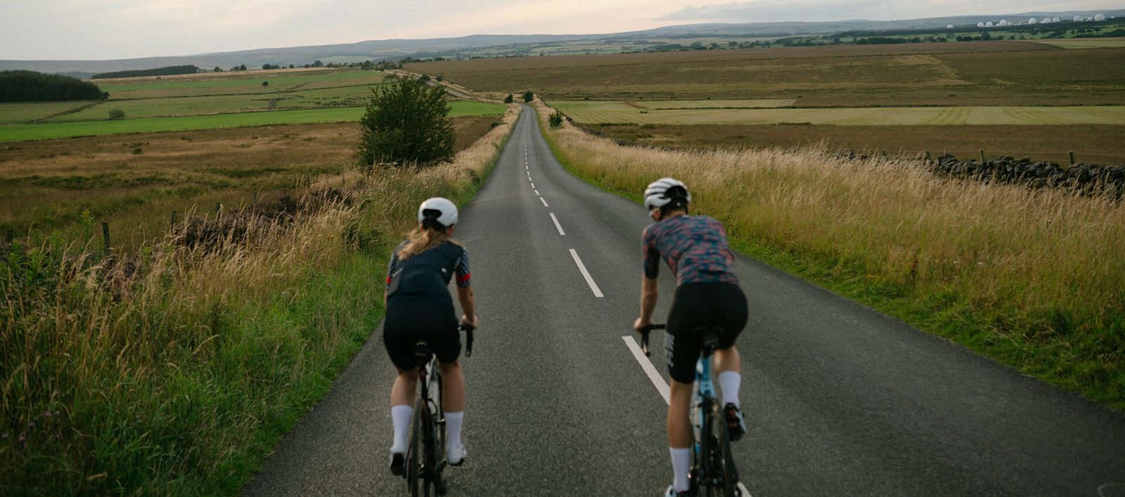 Two cyclists ride on a narrow, paved country road flanked by grassy fields and distant hills. They are wearing cycling gear and helmets as they pedal away from the camera, moving towards the horizon under a partly cloudy sky.